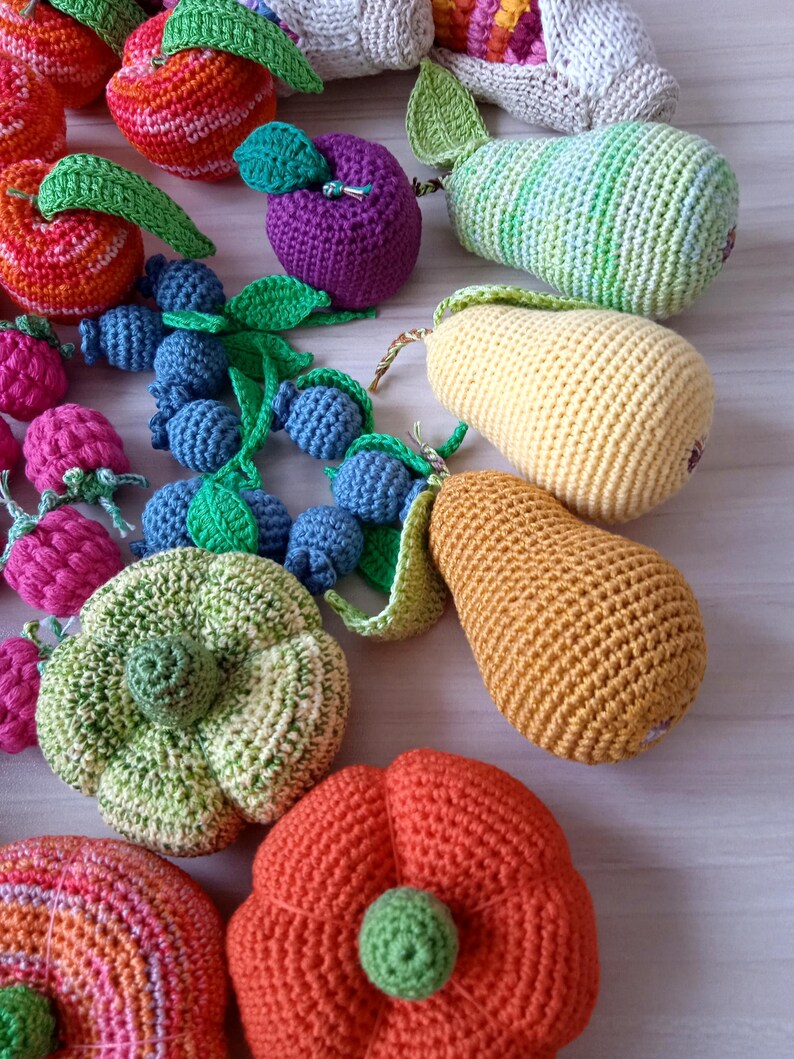 May include: A collection of crocheted fruit and vegetables, including a red apple, a purple plum, a green pear, a yellow pear, a green zucchini, a red pumpkin, and a cluster of blueberries.