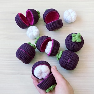 May include: Crocheted mangosteen fruit toys. The purple outer shell is split open to reveal a white interior. Some are whole, others are halved, and a few are open to show the white fruit inside. Green crocheted leaves are attached to the top of the fruit.