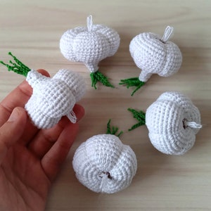 May include: Five crocheted garlic bulbs with green stems. The garlic bulbs are white and have a textured surface.