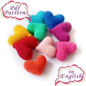 Può includere: Un set di dieci cuori all'uncinetto in vari colori, tra cui rosso, rosa, giallo, verde, blu e arancione. I cuori sono disposti in cerchio, con il testo "Pdf Pattern" in un fumetto sopra e "in English" in un fumetto sotto.