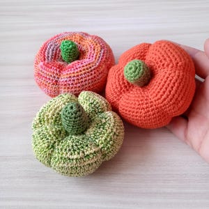 May include: Three crocheted pumpkins in different colors: pink, orange, and green. The pumpkins are all about the same size and have a small green stem on top.