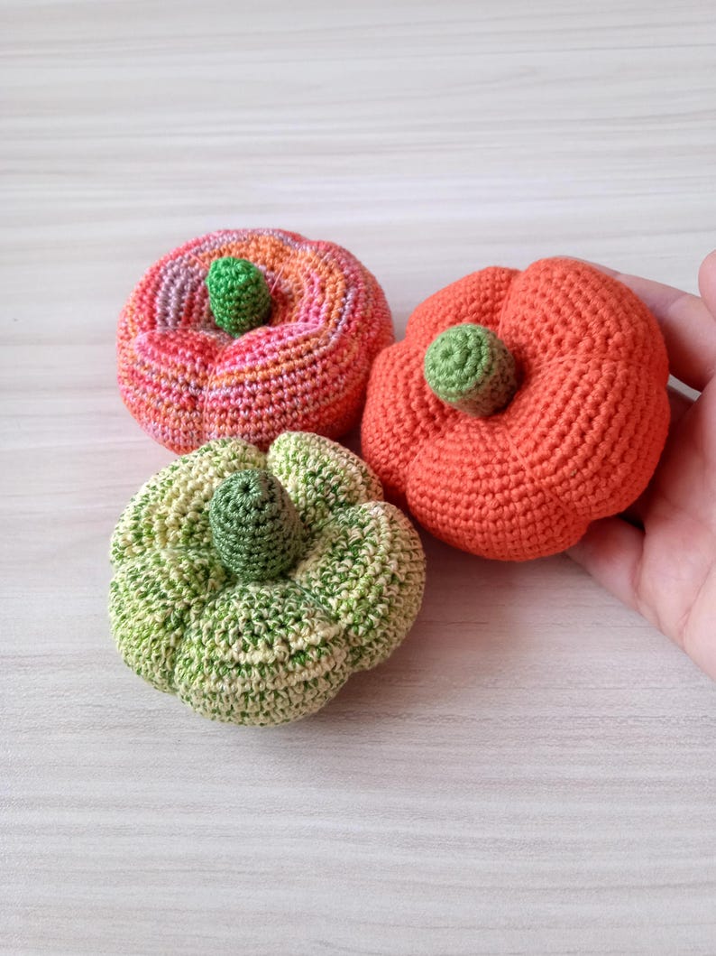May include: Three crocheted pumpkins in different colors: pink, orange, and green. The pumpkins are all about the same size and have a small green stem on top.