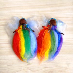 May include: Two handmade felt angel ornaments. Each angel has a brown head, blue beaded halo, and rainbow-colored skirt. White fluffy wings extend from the back. The angels are displayed on a light wood surface.