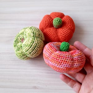 May include: Three crocheted pumpkins in different colors and patterns. One is green and white, one is orange and white, and one is orange and pink.
