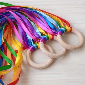May include: Rainbow ribbon toy with three wooden rings. The ribbons are tied together at the top and hang down in a cascade of colours.