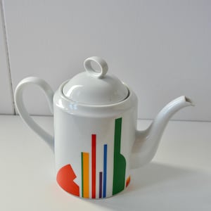 May include: A white ceramic teapot featuring a cylindrical body and a matching lid with a loop handle. The exterior is decorated with a series of vertical lines in red, yellow, orange, blue, and purple, alongside a solid green shape and a red arc.