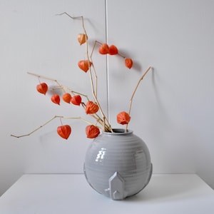 May include: A gray ceramic vase with a round shape and a textured surface, holding dried branches with vibrant orange, heart-shaped flowers. The vase sits on a white surface against a white wall.