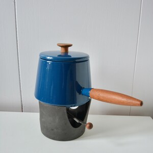May include: A vintage fondue pot. The cobalt blue enamel pot has a wooden handle and knob. It sits on a dark grey metal base with a built-in burner. A classic piece for entertaining, perfect for dinner parties.