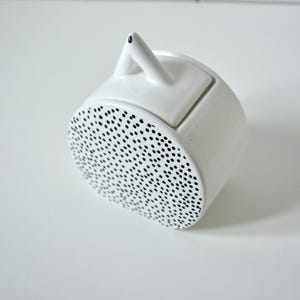 May include: A white ceramic container with a cylindrical body and a lid featuring a small triangular handle. The front circular face is decorated with a dense pattern of small black dots, while the rest of the object remains solid white.