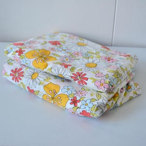 May include: Two folded pieces of floral fabric. The fabric has a white base with a repeating pattern of large yellow, red, pink, and blue flowers. The fabric is neatly folded on a white surface.
