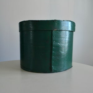 May include: A cylindrical dark green wooden box with a lid. The exterior features a visible wood grain texture and a vertical overlapping seam secured with a series of small metal tacks or nails running from the top edge to the bottom base.