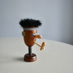 May include: A whimsical wooden figurine with a dark brown body, black fur hair, and a long, wooden nose. The figurine has painted blue and red details on the face and a wooden leg. It stands on a dark brown base.
