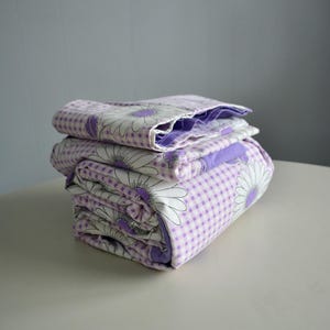 May include: Folded bedding with a daisy floral design. The reverse side has a white and purple gingham check pattern. The edges show a solid purple fabric. The bedding is neatly stacked.