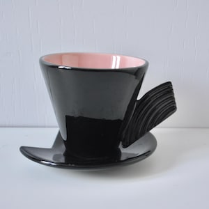 May include: A black ceramic teacup and saucer set with a glossy finish. The interior of the cup is pink. The cup has a unique handle design with a series of curved lines. The saucer is a flat, circular base.