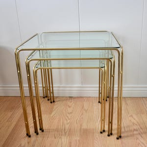 May include: Set of three nesting tables with square glass tops and gold-colored metal frames. The tables are arranged in a nested configuration, with the smallest table fitting under the medium-sized table, and the medium-sized table fitting under the largest table.