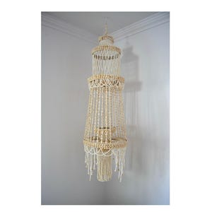 May include: A hanging, tiered chandelier made of natural seashells and off-white string. The chandelier has a cylindrical shape with multiple levels, each adorned with shells and dangling strands. The design is intricate and decorative, suitable for coastal or bohemian decor.