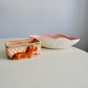May include: A rectangular ceramic planter featuring a relief of a brown dog on the front with a diamond-patterned side, paired with a shallow, oval-shaped ceramic bowl that has a smooth pink interior and a cream-coloured exterior with dark lines.