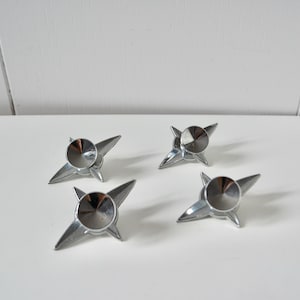 May include: Four shiny, silver starburst-shaped cabinet knobs with a circular center. The knobs are arranged on a white surface, each with four pointed arms extending from a central, domed disc.
