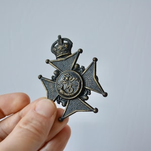 May include: An antique bronze cross-shaped medal with a crown. The medal has a central circular emblem with ornate detailing. The design features a dark bronze colour with gold accents, creating a vintage aesthetic. A decorative item.
