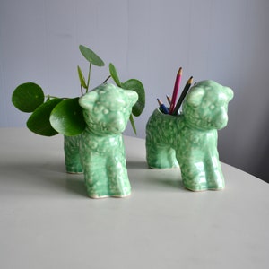 May include: Two light green ceramic sheep-shaped planters. One holds a leafy green plant, the other holds coloured pencils. The planters have a textured surface and are on a white table. The background is a light grey wall.