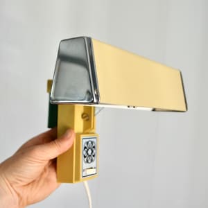 May include: A vintage, rectangular desk lamp with a chrome top and a light yellow shade. The lamp has a yellow base with a floral design on the switch and a white cord. The lamp is held by a person.