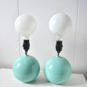 May include: A pair of table lamps featuring smooth, spherical, light mint green ceramic bases. Each lamp has a black socket extending from the top of the sphere, topped with a large, round, white frosted glass globe light bulb.