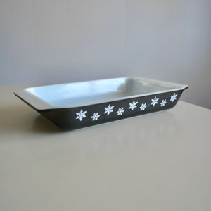 May include: Rectangular baking dish with a white interior and a dark gray exterior. The dark gray exterior is decorated with a row of white snowflake designs. The dish is likely made of ceramic or glass.