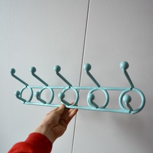 May include: A light blue metal coat rack with six hooks. The rack has a horizontal bar with six circular hooks and six ball-shaped tops. The rack is held by a person wearing a red sweater.