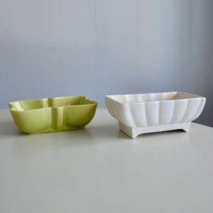 May include: Two ceramic planters. One is a light green, three-sectioned dish with a scalloped edge. The other is a white, rectangular planter with a fluted design and a pedestal base. Both are on a white surface.