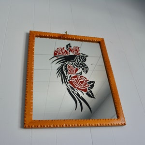 May include: A rectangular wall mirror with a light brown wooden frame featuring a notched edge. The glass surface displays a black graphic of a bird with long tail feathers surrounded by three red roses with black outlines.