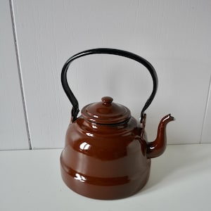 May include: A glossy brown enamel tea kettle with a rounded body, a matching lid with a small knob, and a curved spout. The kettle features a black metal arched handle attached to the sides, providing a contrast to the solid brown finish.