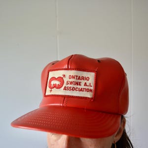 Può includere: Un cappellino da baseball rosso con una toppa rettangolare bianca sul davanti. La toppa mostra la sagoma di un maiale ricamata in rosso, simboli maschili e femminili e il testo rosso "ONTARIO SWINE A.I. ASSOCIATION" in grassetto.