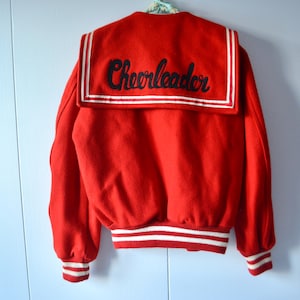 May include: A red wool jacket with a sailor collar and white trim. The word "Cheerleader" is embroidered in black script across the collar. The cuffs and waistband have white stripes.