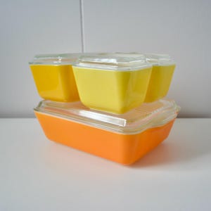 May include: A collection of vintage Pyrex food storage containers. The set comprises a large orange rectangular dish with a clear lid, and three smaller yellow square containers with clear lids. The containers are stacked on a white surface.