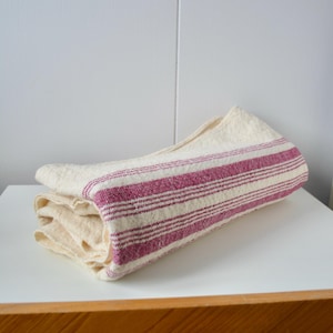 May include: A folded rectangular textile piece in an off-white cream colour featuring a series of parallel magenta pink stripes running along the length of the fabric. The material has a visible woven texture with a slightly coarse appearance.