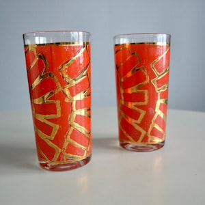 May include: Two tall, clear drinking glasses with a vibrant orange background and a gold geometric design. The rims of the glasses are also trimmed with gold. The glasses are set on a white surface.