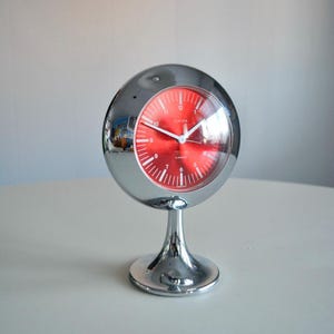 May include: A chrome-finished, spherical table clock with a vibrant red face. The clock features white hour markers and hands, and is mounted on a pedestal base. The clock's face reads "Europa".
