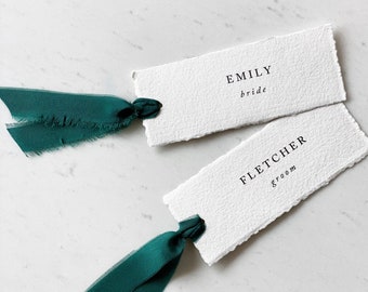 May include: Two white textured place cards with the names "Emily, bride" and "Fletcher, groom" printed in black. Each card is tied with a dark green ribbon. The cards are on a white marble surface.