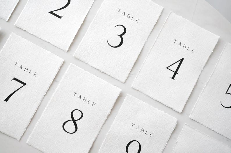 Op de afbeelding: Witte rechthoekige tafelnummerkaarten met zwarte tekst. Elke kaart heeft een uniek nummer van 2 tot en met 9, met het woord "TABLE" boven het nummer. De kaarten hebben een textuur, handgemaakte papieruitstraling.