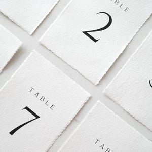 May include: White table number cards with black numerals and the word "TABLE" printed at the top. The cards have a textured, deckled edge. Numbers 1, 2, 7, and 8 are visible. Ideal for weddings or events.