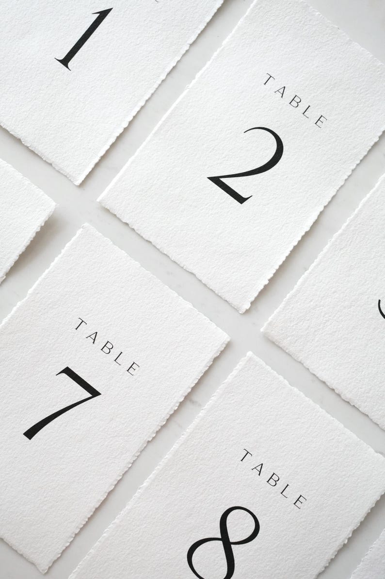 Op de afbeelding: Witte tafelnummerkaarten met zwarte cijfers en het woord 'TABLE' bovenaan gedrukt. De kaarten hebben een getextureerde, gescheurde rand. Nummers 1, 2, 7 en 8 zijn zichtbaar. Ideaal voor bruiloften of evenementen.