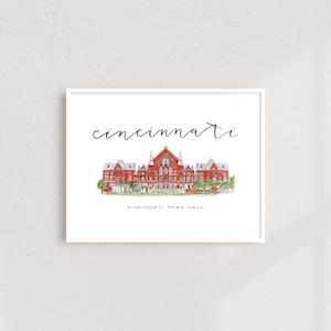 May include: Watercolor print of Cincinnati Music Hall in a white frame. The word "cincinnati" is written above the building in cursive. The building is red brick with a detailed architectural design, and green trees are in front.