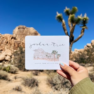 May include: A white card with the words "joshua tree" in cursive and a watercolor illustration of the Joshua Tree National Park. The card is held up against a backdrop of the park's landscape and a clear blue sky.