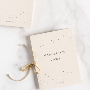 May include: Two cream-colored vow books with gold ribbon. One book reads "Tyler's Vows" and the other reads "Madeline's Vows." Both books have a scattering of small stars and the date 07.17.21 printed on them. The books are on a white surface.