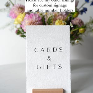 Op de afbeelding: Een wit papieren bord met de zwarte tekst "CARDS & GIFTS", gehouden door een klein houten blok. De achtergrond toont een boeket van roze, gele en blauwe bloemen.