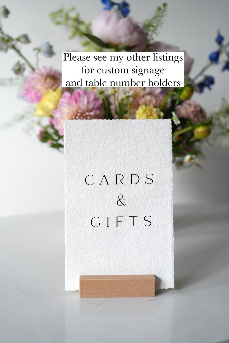 Op de afbeelding: Een wit papieren bord met de zwarte tekst "CARDS & GIFTS", gehouden door een klein houten blok. De achtergrond toont een boeket van roze, gele en blauwe bloemen.