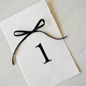 May include: A white textured paper table number card with a black number 1 and a black ribbon bow. The card has a deckled edge and is angled on a white marble surface. Ideal for weddings or special events.
