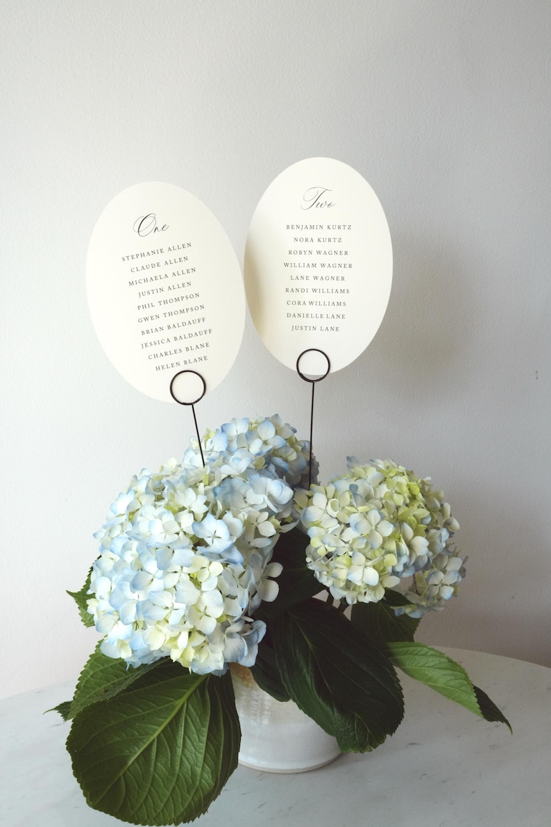 Op de afbeelding: Twee ovale, gebroken witte tafelkaarten met de tekst One en Two en zwart gedrukte gastennamen, elk gehouden door een dunne zwarte draadstandaard in een witte vaas met blauwe en groene hortensia's en grote groene bladeren.