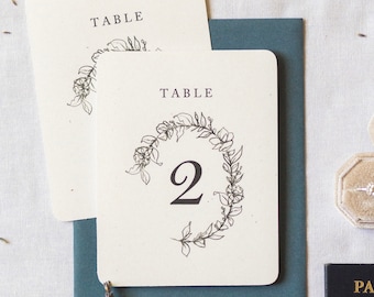 May include: Cream-colored table number cards with rounded corners, featuring a floral wreath design and the number 2. The cards are placed on a teal envelope. Additional stationery includes a name card and a small black book.