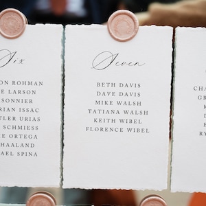 May include: Close-up of elegant wedding seating cards. Each card is white with a deckled edge, featuring names in black script. A rose-gold wax seal adorns each card. The cards are numbered with the words "Six", "Seven", "Eleven", and "Twelve".